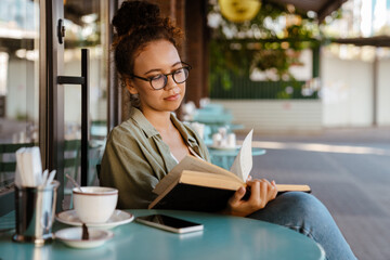 White young woman drinking coffee while reading book in cafe outdoors