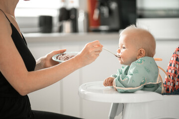 Mother spoon feeding her baby boy child in baby chair with fruit puree in kitchen at home. Baby solid food introduction concept.