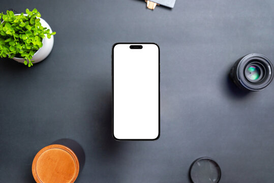Phone with isolated transparent display floats above the desk. The front camera is built into the display. Modern round and thin edges. Black table with plant, lens and box. Top view, flat lay