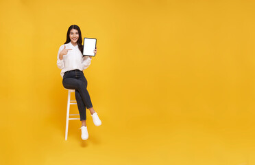 Fototapeta na wymiar Happy young asian woman showing empty tablet screen while sitting on white chair on yellow studio background. full body length. Excited lady recommending nice and useful tablet application
