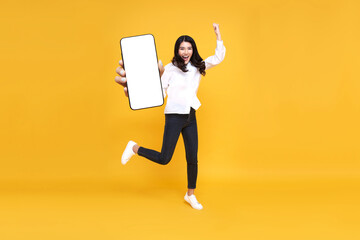 Happy young asian woman showing empty mobile phone screen while jumping up on yellow studio background. full body length. Excited lady recommending nice and useful mobile application