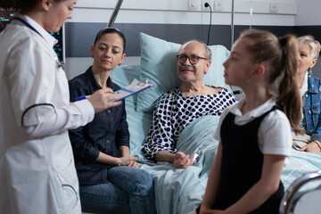 Medical professional performing vital signs check to sick elderly man resting in geriatric hospital bed. Little girl asking female doctor about health condition of hospitalized grandfather.