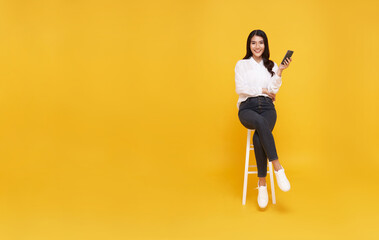 Happy young Asian smile face woman using mobile phone. While her sitting on white chair and looking on yellow copy space background.