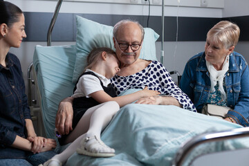 Elderly man in geriatric clinic bedroom hugging little girl. Senior patient relatives visiting medical tower recovery room. Granddaughter visiting sick grandfather at sanatorium. - obrazy, fototapety, plakaty