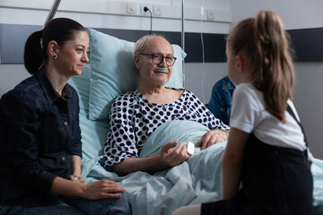 Elderly man lying in hospital bed chatting with visiting family members. Little girl talking to grandfather in elderly people medical clinic. Family living with old man resting in sanatorium bed. - obrazy, fototapety, plakaty