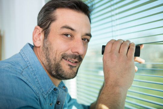 excited man spying through the window blinds with binoculars
