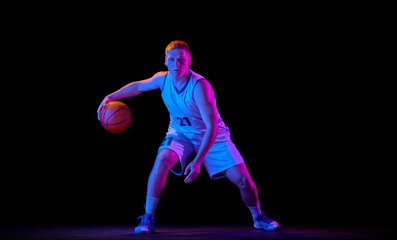Fototapeta premium Dynamic portrait of young active athlete, male basketball player in sports uniform practicing with ball, jumping, dribbling isolated over dark background in neon light