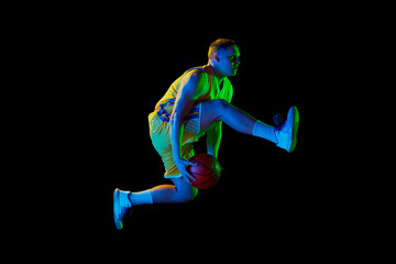 Fototapeta na wymiar Studio shot of young active athlete, male basketball player in sports uniform in motion and action with ball isolated over dark background in neon light