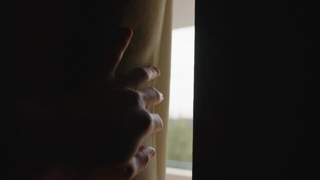 Hand slowly moves and open heavy window curtain letting sunlight in dark room. In complete darkness male hand push a fabric curtain as light enters the room to reveal the sunny day outside with trees