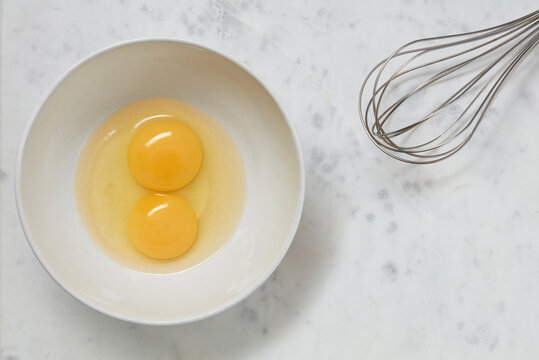 Double yolk eggs in a white bowl and wire whisk on a white marble surface. Two yolks in one chicken egg. Identical twins. Double eggs. Flat lay