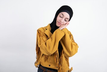 Relax and sleep time. Tired young beautiful muslim woman wearing hijab and yellow jacket over white background with closed eyes leaning on palms making sleeping gesture.