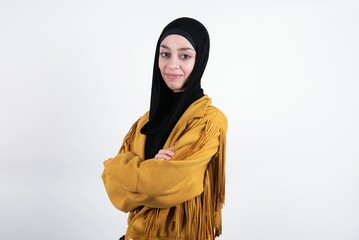 Image of cheerful young beautiful muslim woman wearing hijab and yellow jacket over white background with arms crossed. Looking and smiling at the camera. Confidence concept.