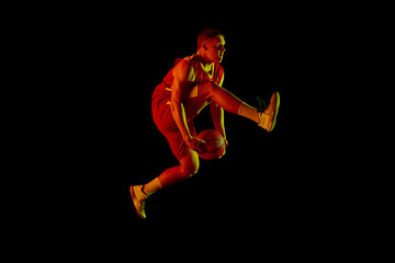 Dynamic portrait of young active athlete, male basketball player in sports uniform practicing with ball, jumping, dribbling isolated over dark background in neon light