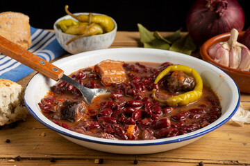 Close-up of Basque bean stew with spoon on rustic table with onions, garlic, green chilli and bread, horizontal