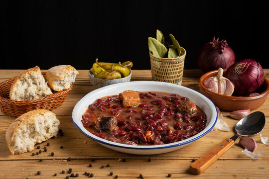 View of a plate with Basque bean stew on a rustic table with onions, garlic, green chillies and bread, black background, horizontal