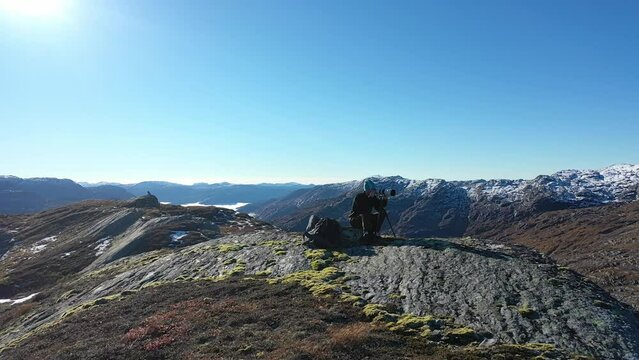 Landscape photographer with telezoom and tripod on tall mountain peak during fall season - Gentle orbit around person and in front of lens with sunshine and stunning landscape all around - Norway