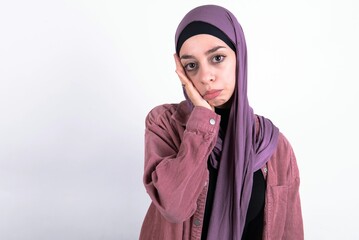 Sad lonely young beautiful muslim woman wearing hijab and pink jacket over white background touches cheek with hand bites lower lip and gazes with displeasure. Bad emotions