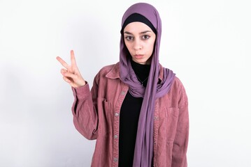 young beautiful muslim woman wearing hijab against white background makes peace gesture keeps lips folded shows v sign. Body language concept