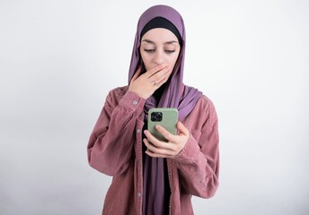 young beautiful muslim woman wearing hijab against white background being deeply surprised, stares...