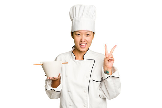 Young chef asian woman holding a bowl of ramen isolated joyful and carefree showing a peace symbol with fingers.