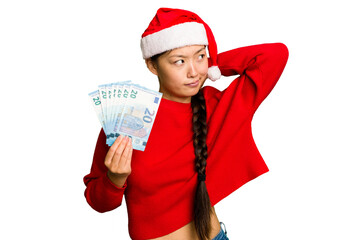 Young asian woman celebrating Christmas holding banknotes isolated touching back of head, thinking and making a choice.