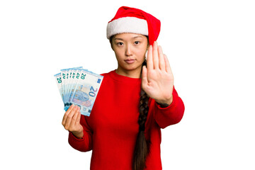 Young asian woman celebrating Christmas holding banknotes isolated standing with outstretched hand showing stop sign, preventing you.