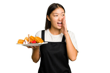 Young asian waitress woman holding waffles isolated shouting and holding palm near opened mouth.