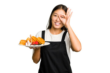 Young asian waitress woman holding waffles isolated excited keeping ok gesture on eye.