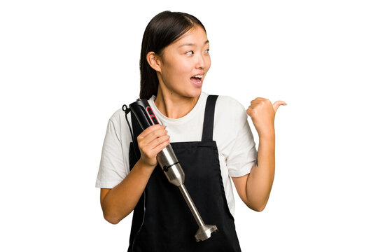 Young asian cook woman holding a blender isolated points with thumb finger away, laughing and carefree.