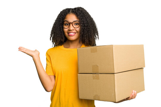 Young african american woman moving to other house while picking up a box full of things isolated showing a copy space on a palm and holding another hand on waist.