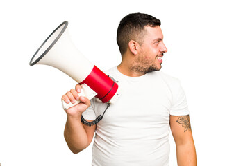 Young caucasian man holding a megaphone isolated looks aside smiling, cheerful and pleasant.