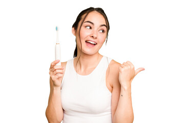 Young caucasian woman holding electric toothbrush isolated on green chroma background points with thumb finger away, laughing and carefree.