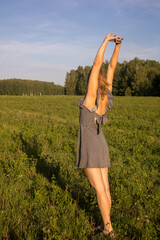a woman in a black sundress with her hair down stands with her back in a field of clover with her hands raised up against a background of blue sky and grass