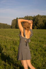 a woman in a black sundress with her hair down stands with her back in a field of clover with her hands behind her head against a background of blue sky and grass
