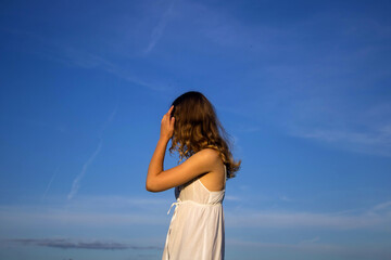 a girl with long curly hair in a white sundress stands with her back against the blue summer sky