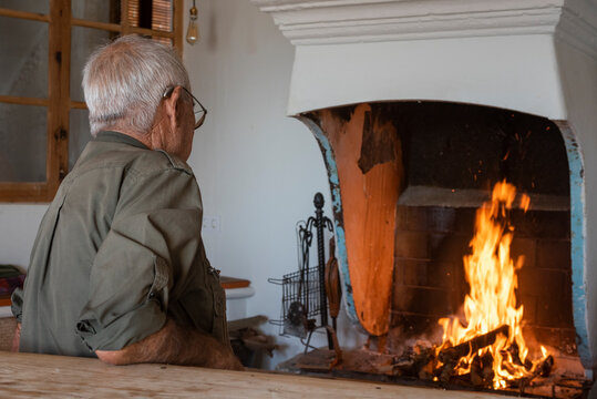 Hombre mirando el fuego en casa rústica vestido con ropa de camuflaje, cazador anciano de la tercera edad blanco y con canas un poco de espaldas