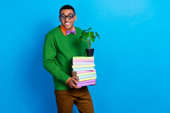 Photo of young cute smiling academic nerd student man wear funky spectacles hold stack books with decorative plant isolated on blue color background
