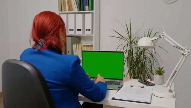 Business woman working at desk looking at green screen laptop and taking notes. Back view of female in blue jacket working remotely.