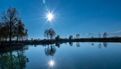 Naklejka na ściany i meble Landscape of lake with sun reflection which is on blue sky, Silhouette of trees in the background