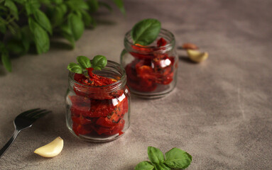dried tomatoes in a glass jar with garlic and basil on a gray background