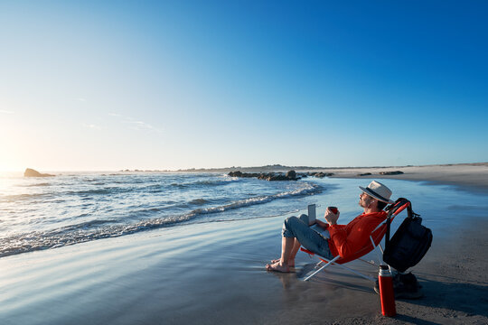 mature latin man sitting on the shore of the beach with his laptop drinking coffee and enjoying