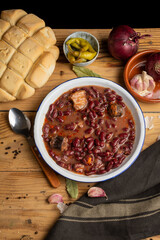 Aerial view of a plate of Basque beans on a rustic wooden table with a spoon, onions, garlic and a loaf of bread, vertical