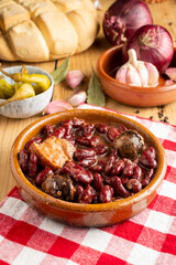 Close-up of red bean stew on red kitchen cloth, rustic wooden table with green chillies, garlic, onions, bay leaf and loaf of bread, selective focus, vertical