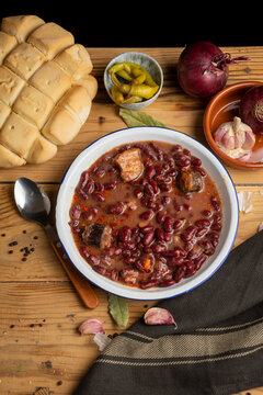 Aerial view of a plate of Basque beans on a rustic wooden table with a spoon, onions, garlic and a loaf of bread, vertical