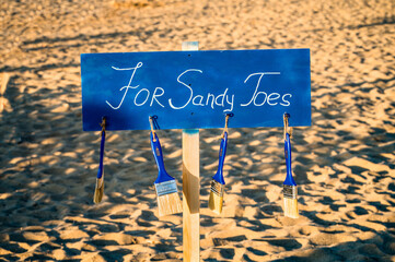On the  beach wooden blue sign, and brushes hanging on it, with signature for sandy shoes. 