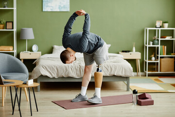 Full length portrait of man with prosthetic leg working out at home and doing stretching exercises,...