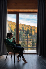 Woman in green sweater sitting with coffee near panoramic window at modern living room with stunning view on autumn forest mountains