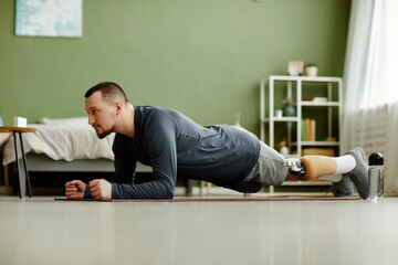 Side view portrait of man with prosthetic leg working out at home and doing plank exercises, copy space