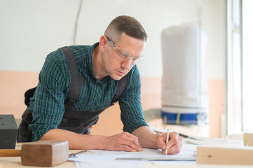 A portrait of a carpenter in goggles and work overalls draws a blueprint for a workshop.