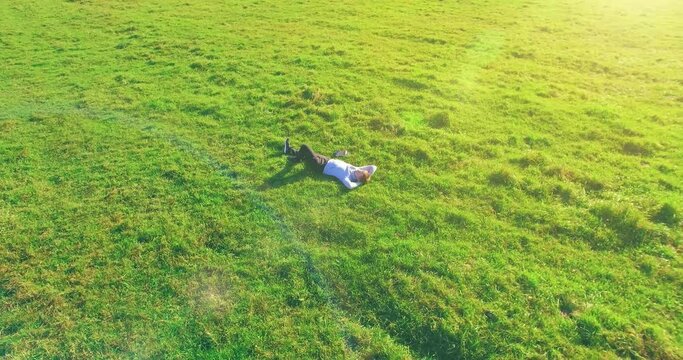 Aerial view point. Low orbital flight around man lied on green grass with notebook (pad) at yellow rural field. Casual weared. Sun rays. Radial movement.